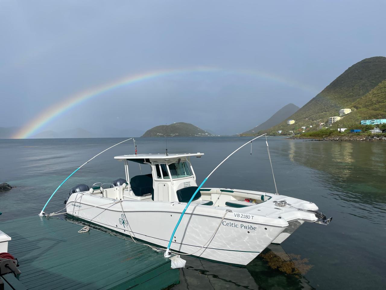 Celtic Pride powerboat docked at the private West End pier in Tortola, BVI, with a rainbow arching over calm waters and green hills in the background.